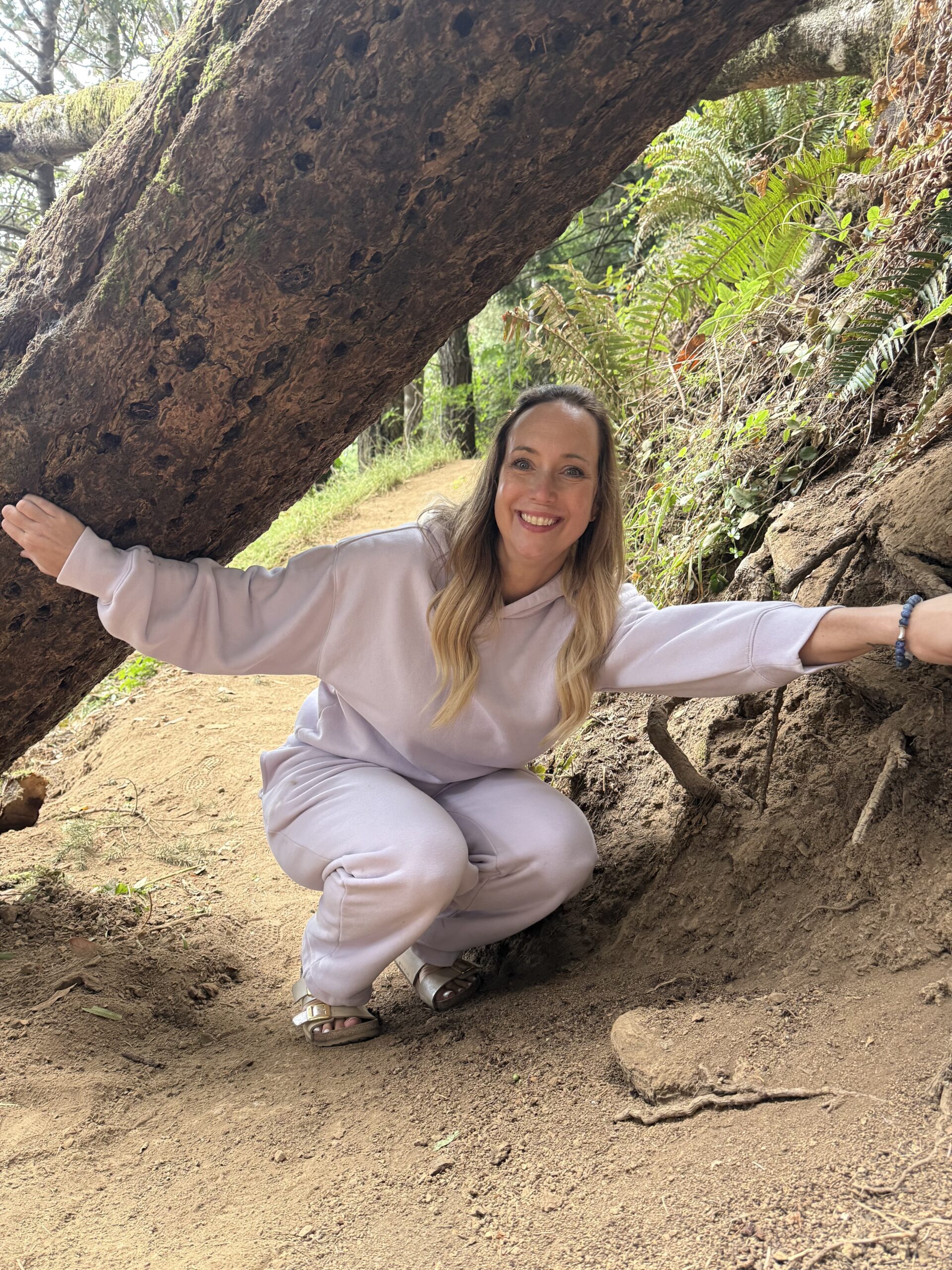 Sara Newman in a lavender lounge set beneath a large tree on a wooded trail